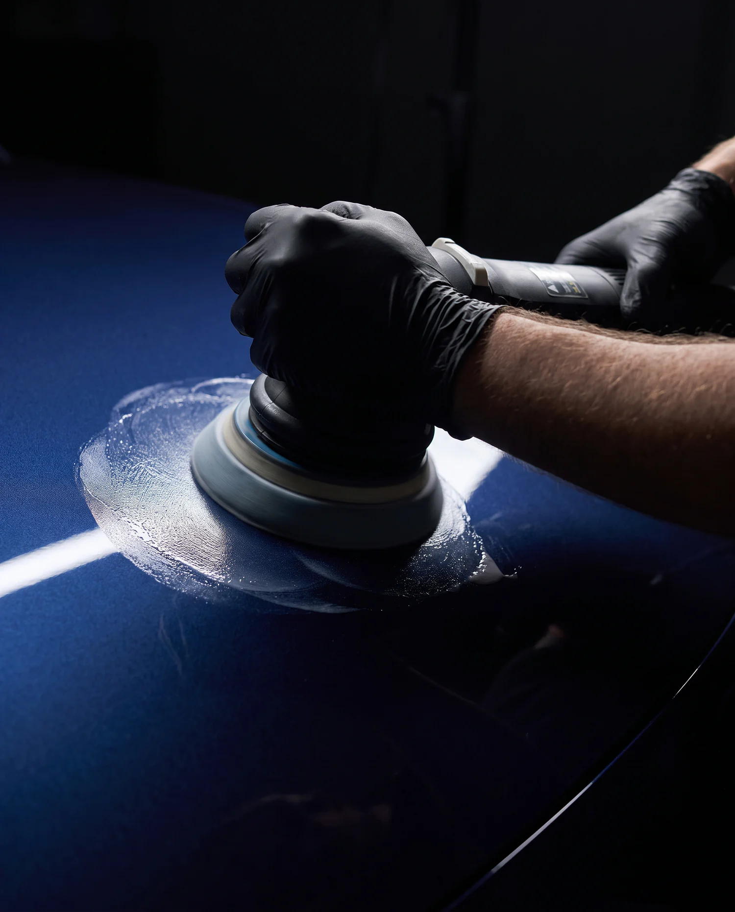 A gloved hand working a dual action polisher across a deep blue car panel under a hard inspection light